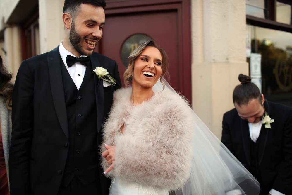 Happy bride and groom in wedding attire standing outside, smiling and looking at each other, with a bridesmaid or guest in the background.