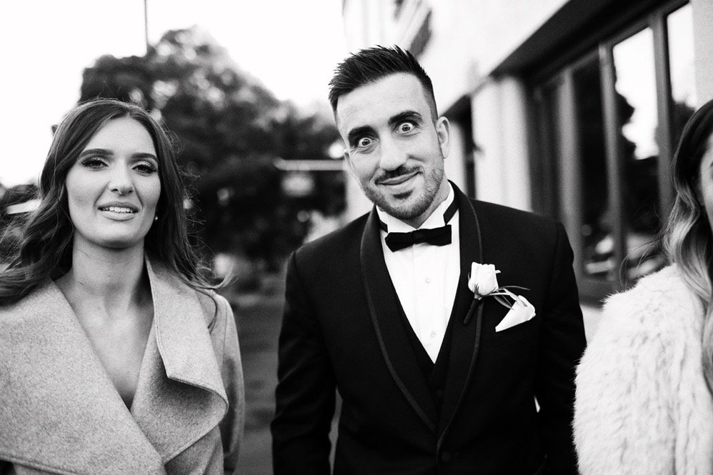 A black and white photo of a man in a tuxedo with a boutonniere, standing outdoors with women nearby, smiling at the camera.