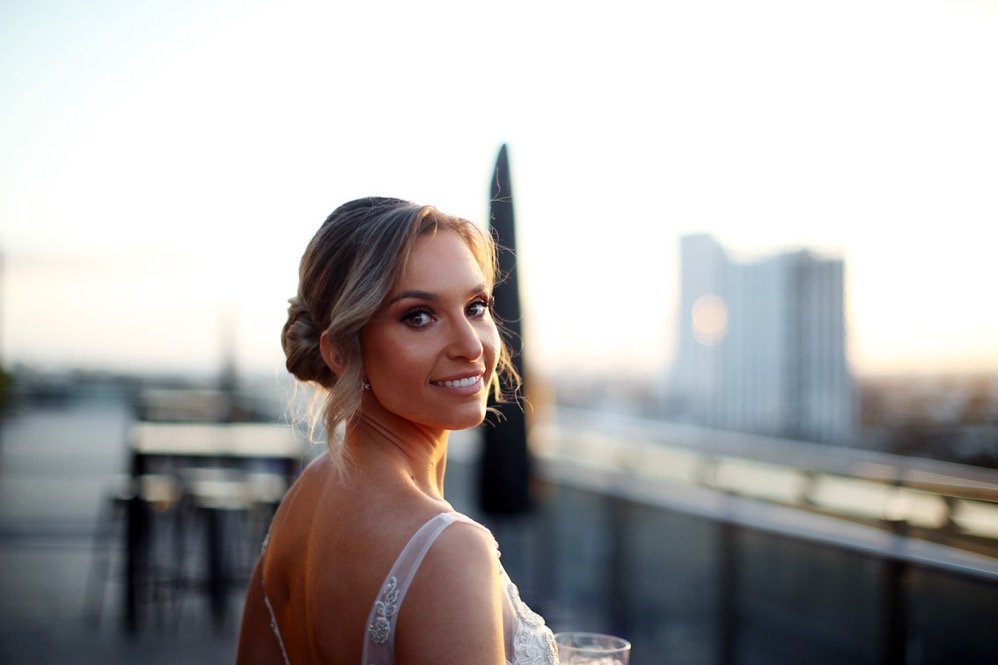 A woman in a white dress with a low back, smiling and holding a glass, on a rooftop at sunset.
