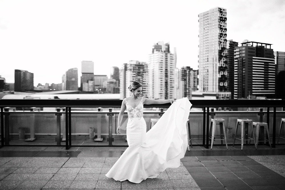 A woman in a wedding dress holding up the train on a rooftop with a city skyline in the background.