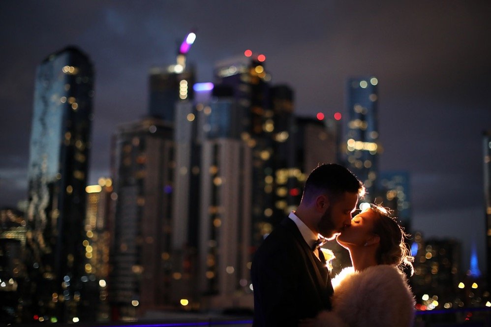 A couple kissing at night against a city skyline with illuminated skyscrapers.