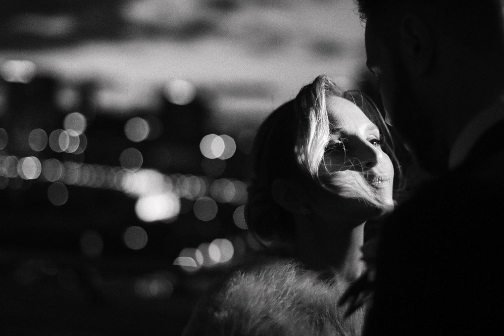 A black and white photo of a woman and a man close together at night, with city lights blurred in the background. The woman is smiling with her eyes closed, and the man is slightly above her, mostly out of focus.
