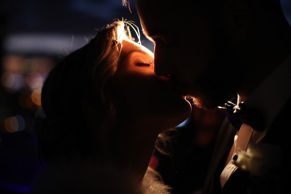 A romantic couple kissing in a dimly lit setting with city lights blurred in the background.