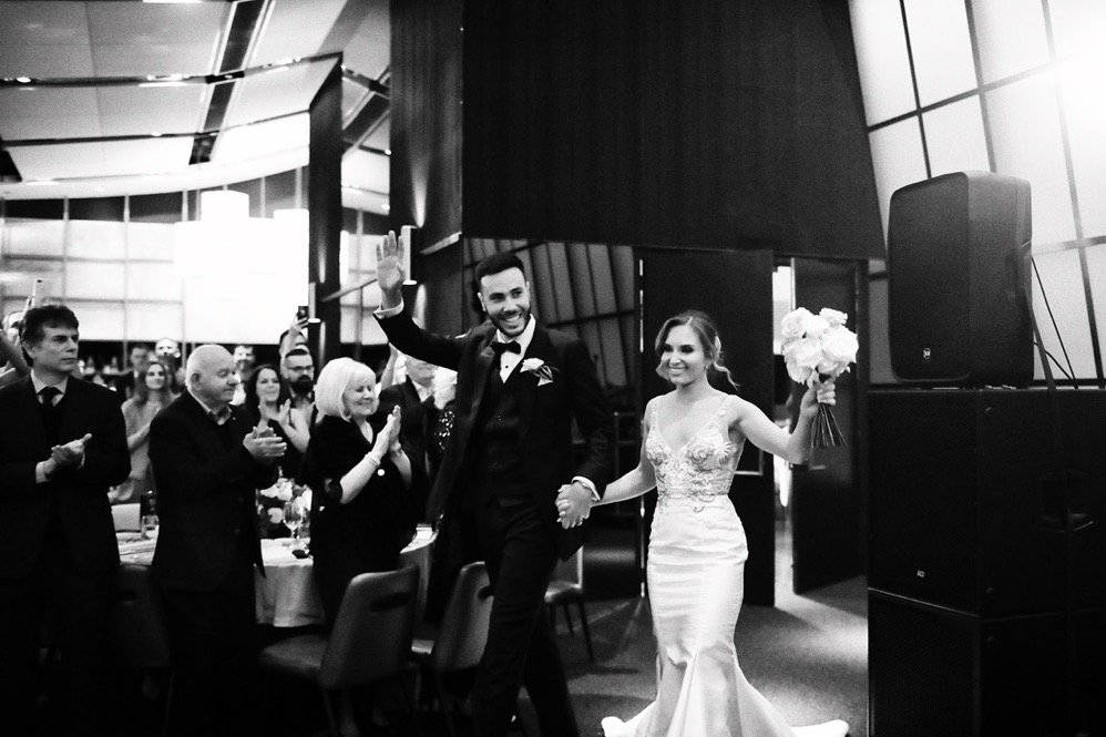 Black and white photo of a newlywed couple walking hand in hand at their wedding reception, surrounded by guests clapping and smiling.
