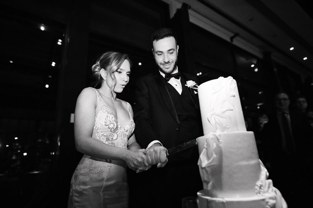 Black and white photo of a bride and groom cutting a wedding cake together at their wedding reception, holding a knife.