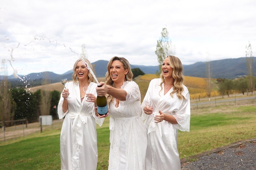 Three women in white robes celebrating outdoors, one popping a champagne bottle with others holding glasses, with mountains and grass in the background.