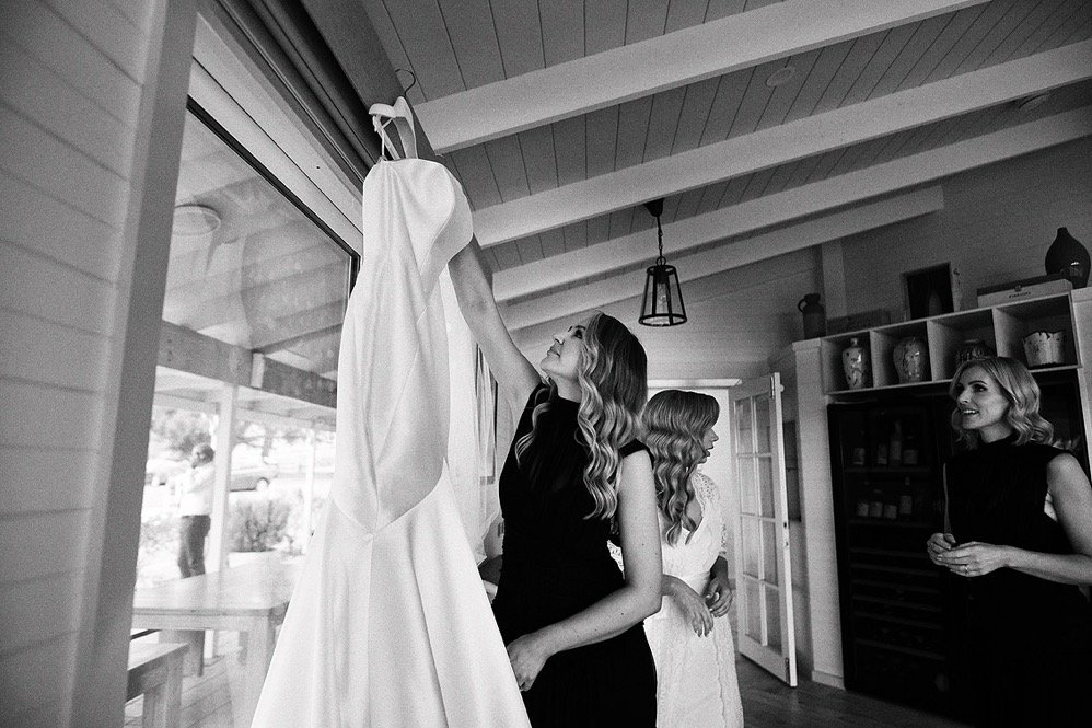 Three women indoors, one holding a wedding dress and examining it, with a window and kitchen cabinets visible in the background.