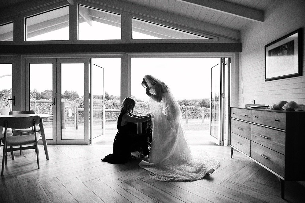 A woman in a wedding dress is bending down to help a girl, likely a flower girl, with her dress inside a modern room with large windows and a view outside.