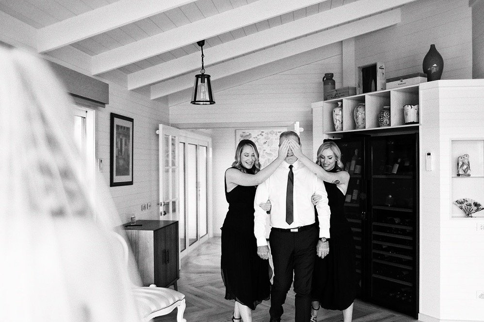 Two women with blonde hair dressed in black dresses are on either side of a man in a white shirt and black tie, covering his eyes with their hands and smiling. The setting is a cozy, well-lit living room with wooden floors, shelves with decorative it