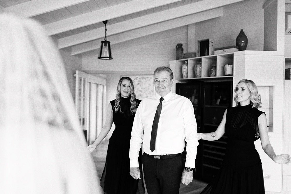 A man in a white shirt and black tie stands between two women in black dresses in a cozy, well-lit room with shelves and decorative items.