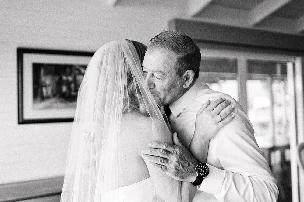 A man and woman sharing a hug, likely on a wedding day, with the man wearing a dress shirt and the woman wearing a veil. The woman is facing away, and the man is kissing her on the cheek, inside a room with windows and framed pictures.
