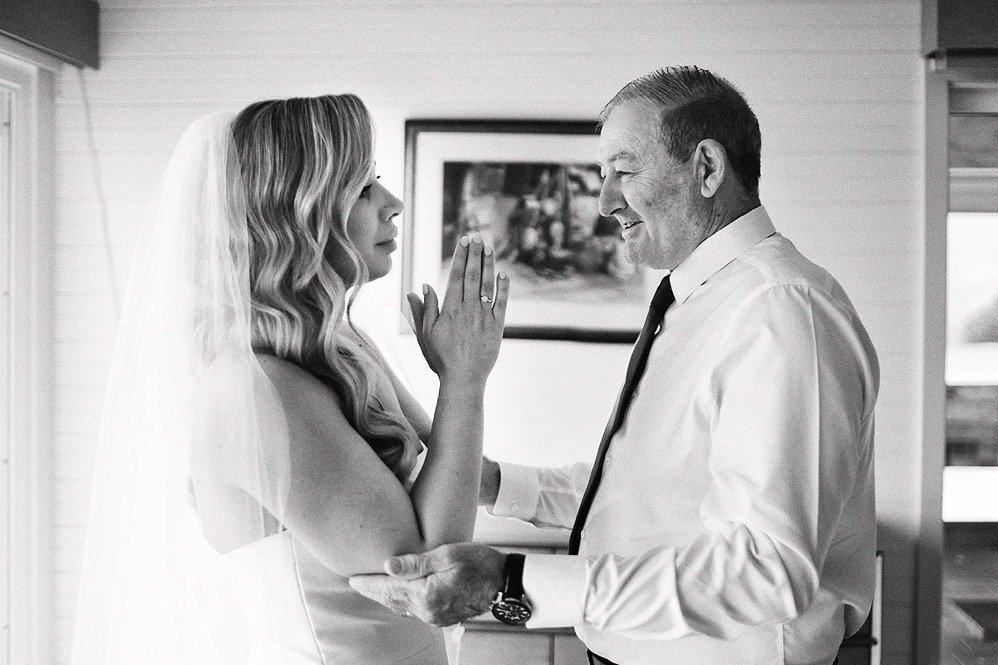 A bride and an older man, possibly her father, sharing a tender moment during her wedding, with the bride touching her forehead to the man's and smiling.