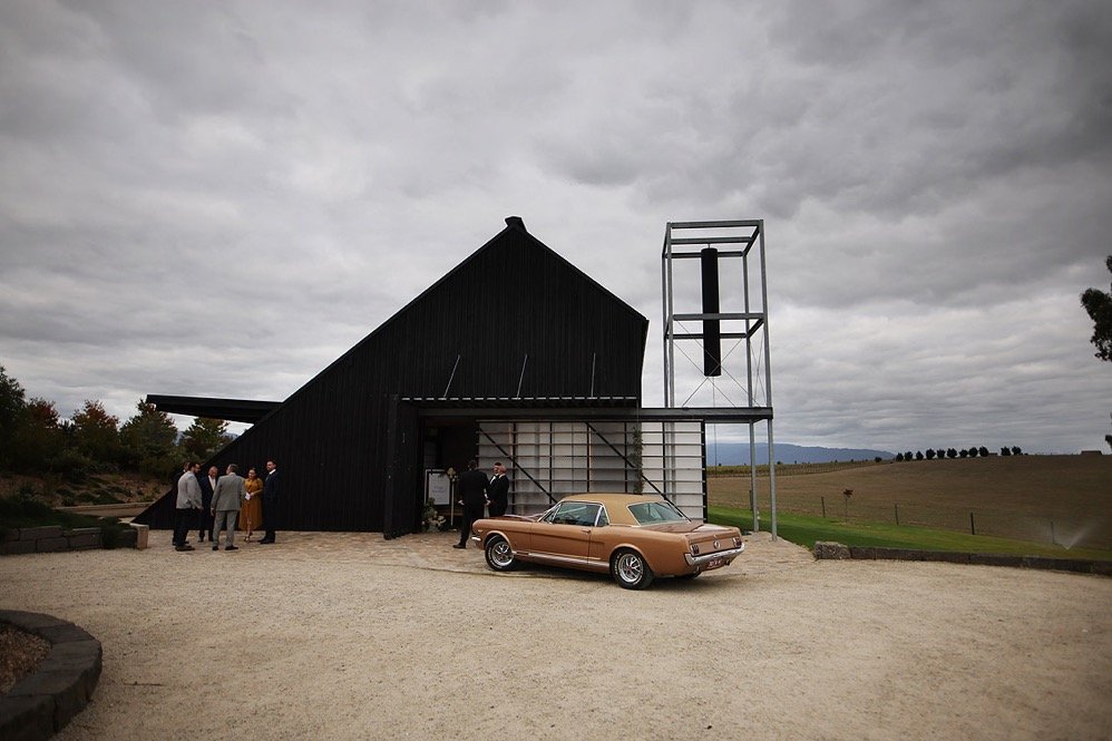 A group of people in formal attire gathered outside a modern black barn with a vintage car parked in front, under a cloudy sky.