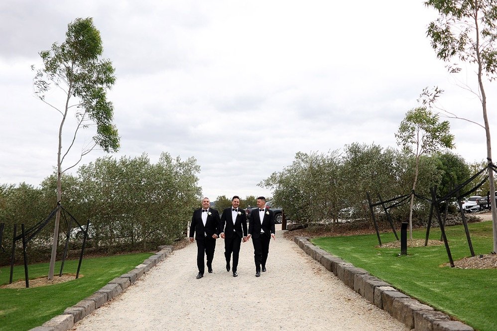 Three men in tuxedos walking down a gravel path surrounded by landscaped grass and trees.