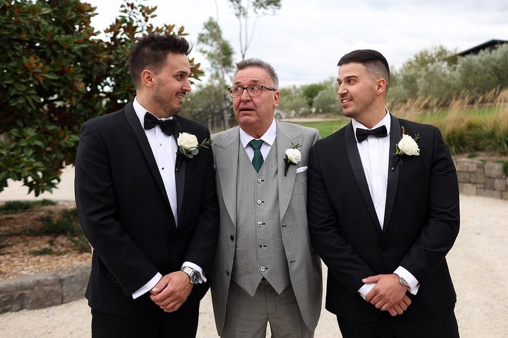 Two men in black tuxedos with white shirts and bow ties, each with a white rose boutonniere, standing on either side of an older man in a gray suit with a light green tie, outdoors during daytime.
