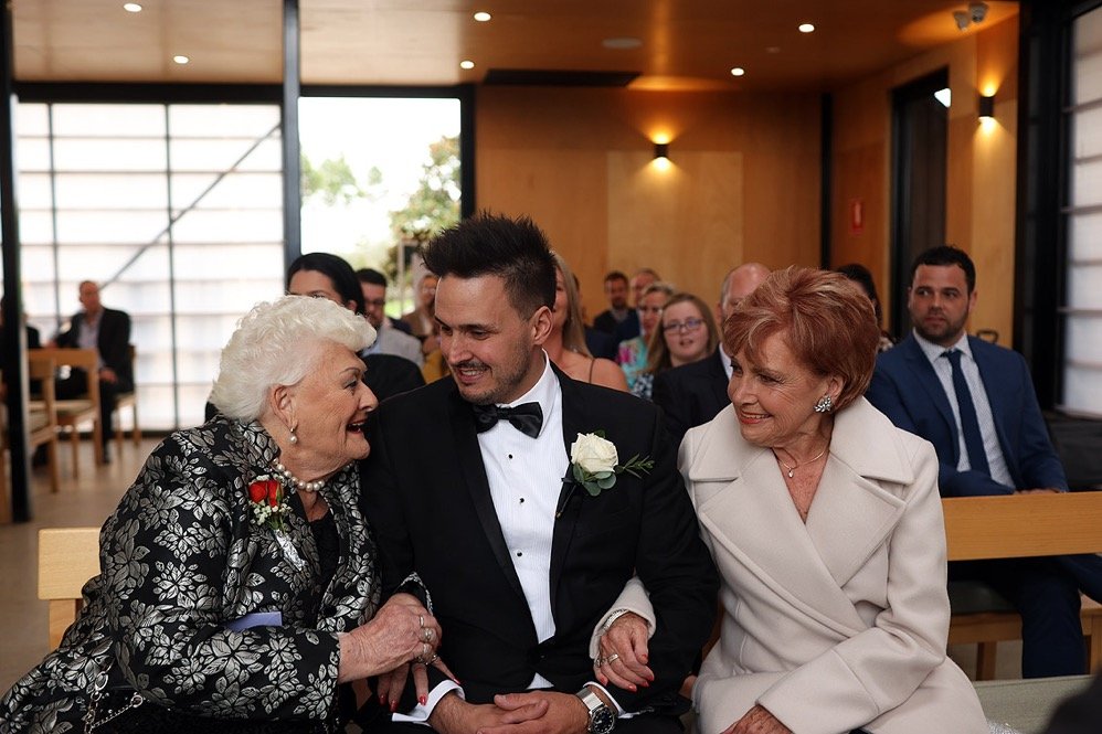 Three people, including a man in a tuxedo, smiling and talking to elderly women at a wedding ceremony, with guests seated in the background.