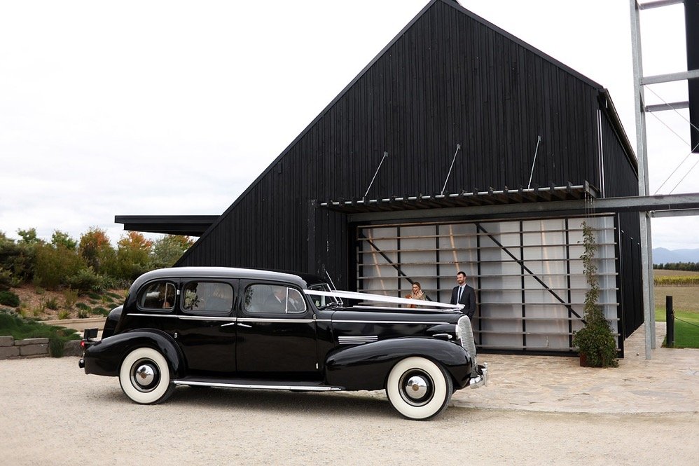 A vintage black car parked outside a modern black building with two people standing in front.