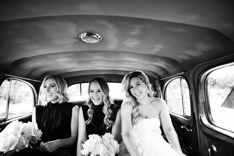 Three women sitting in the backseat of a vintage car, smiling and holding bouquets, in black and white.