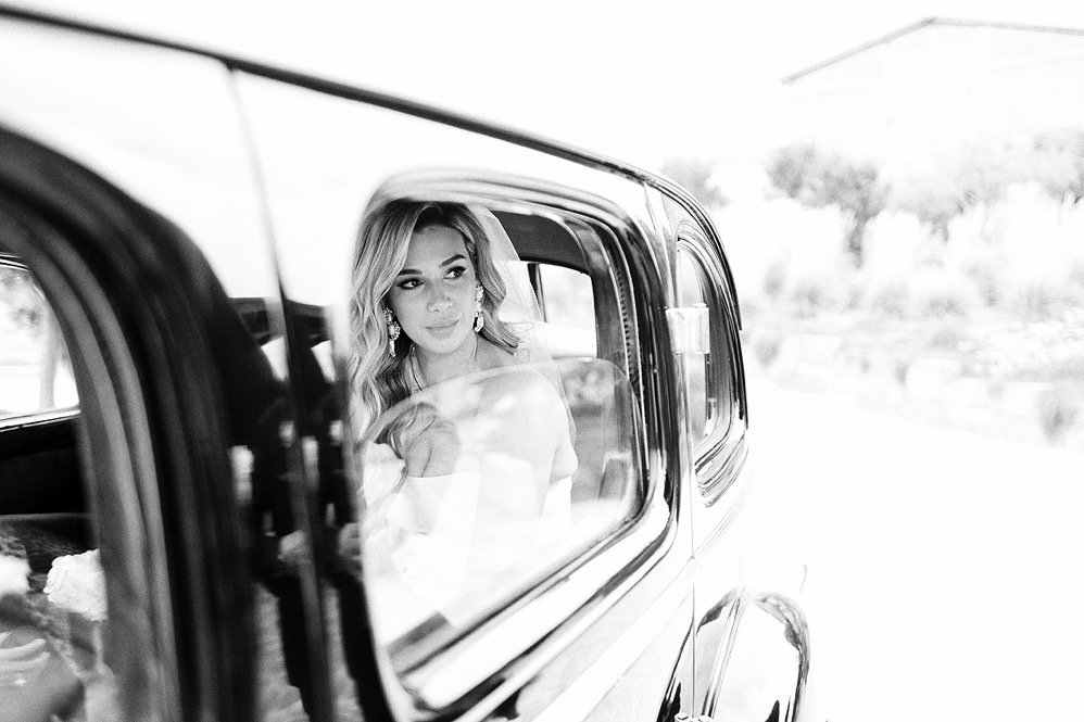A woman with wavy hair and earrings is looking out of the window of a vintage car.