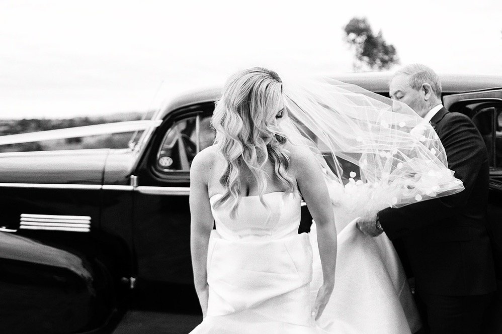 A bride in a white wedding dress with long, wavy hair standing outside near a vintage black car, with a man in a tuxedo holding her veil.