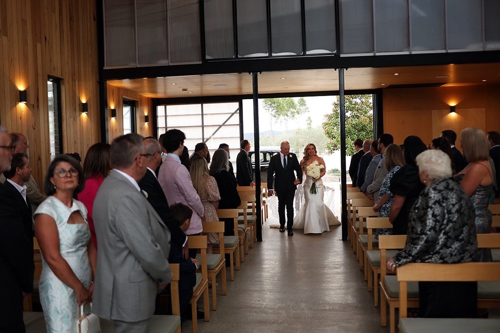 Bride walking down aisle with her father at a wedding ceremony in a modern venue with wooden walls and large windows.