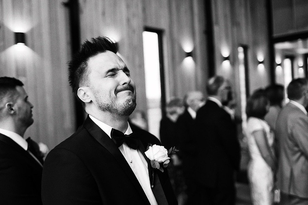 A groom in a tuxedo with a bow tie and a boutonniere, making a facial expression that shows he is emotional at a wedding or formal event, with other guests in the background.