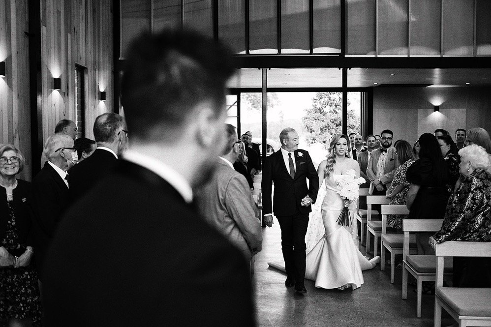 A bride walking down the aisle holding a bouquet, escorted by her father during a wedding ceremony in a modern church or chapel with guests seated on either side.