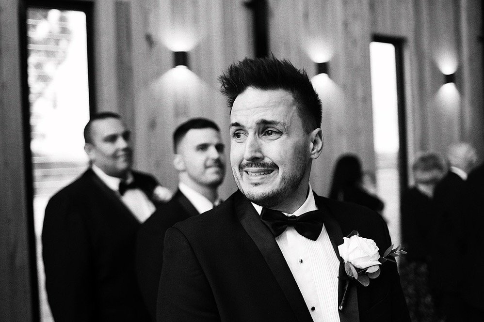 A groom in a tuxedo with a boutonnière looks emotional during a wedding ceremony, with groomsmen in tuxedos in the background.