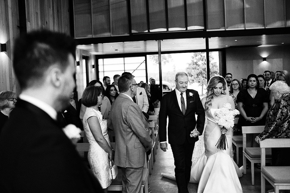 A bride walking down the aisle holding a bouquet of flowers, being escorted by an older man in a suit, during a wedding ceremony inside a modern venue with large windows and seated guests.
