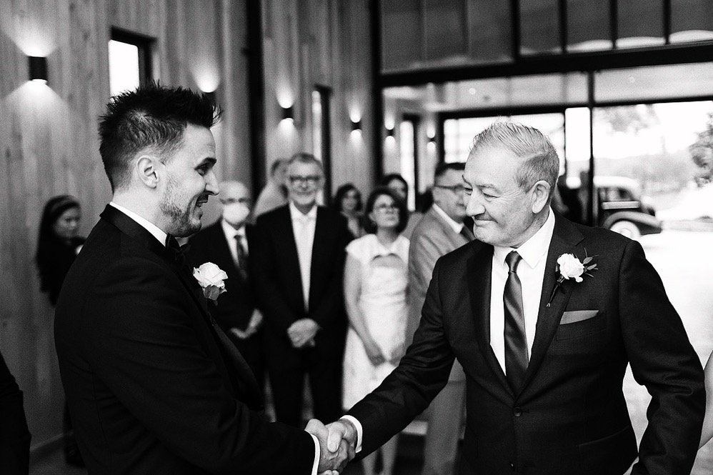 Two men in suits shaking hands during a wedding ceremony, with guests in the background inside a wooden venue.
