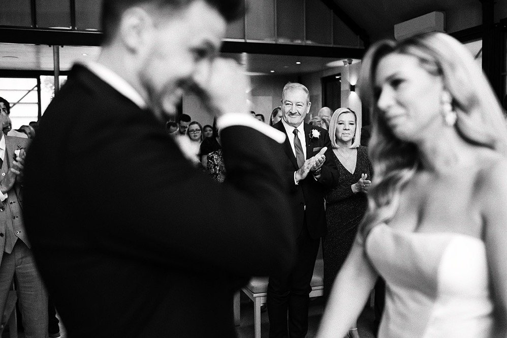 A wedding ceremony scene with a groom, bride, and guests clapping inside a modern venue.