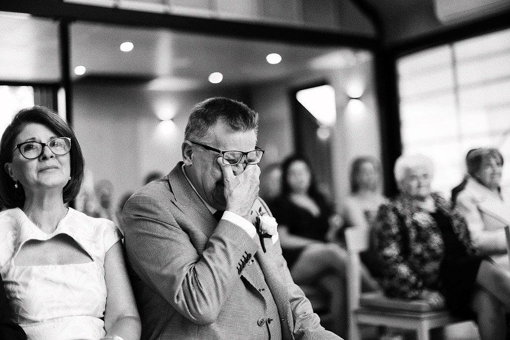 An emotional man in glasses and a suit at a formal event, with his hand covering his face, sitting next to a woman with glasses. Several people seated in the background.