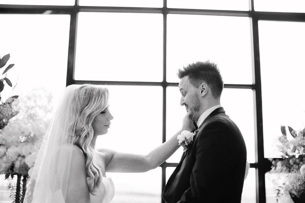 A bride and groom in a wedding ceremony, standing in front of large window panes. The bride reaches out to touch the groom's face.