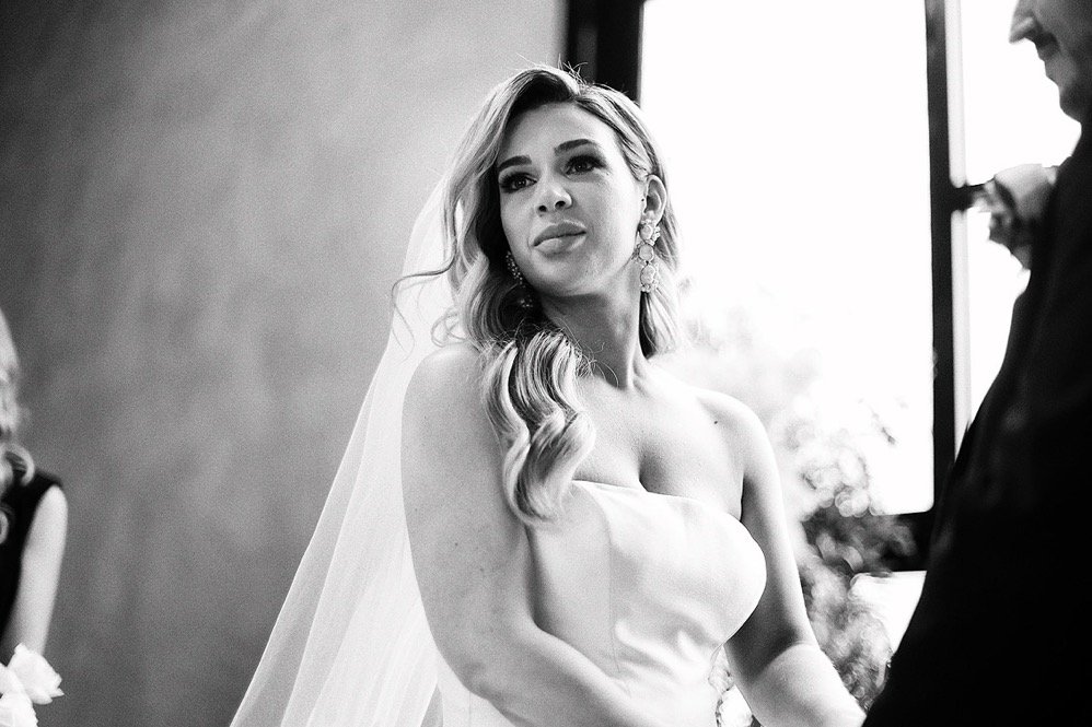 A bride with wavy hair and earrings wearing a strapless wedding dress and veil, standing and looking slightly to the side during a wedding ceremony.