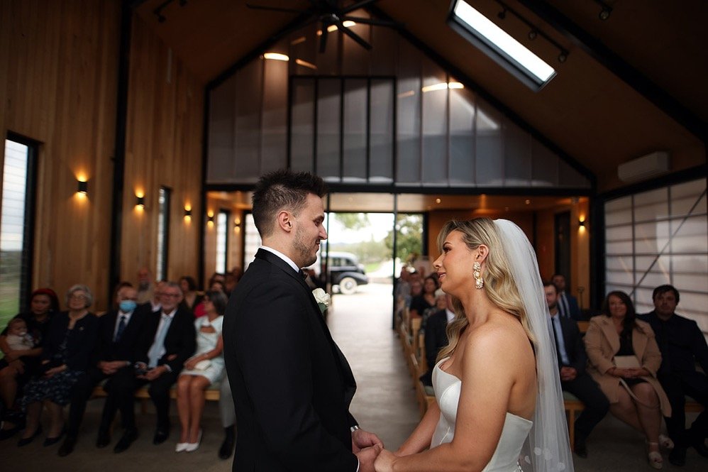 Bride and groom holding hands during their wedding ceremony inside a wooden hall. Guests seated and watching, some wearing masks. Large windows and ceiling with skylight.