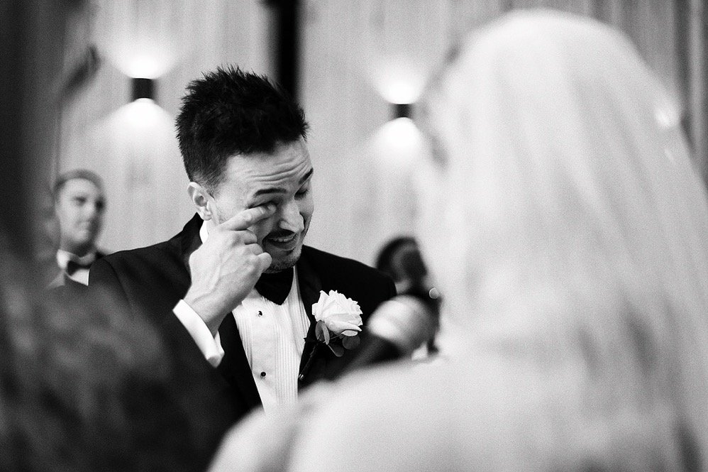 A man in a tuxedo wiping a tear from his eye at a wedding reception, with a woman in a wedding dress and other guests in the background.