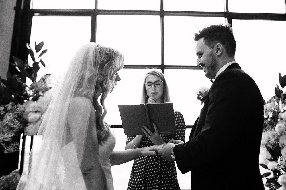 Bride and groom holding hands during a wedding ceremony, with a officiant reading from a book.
