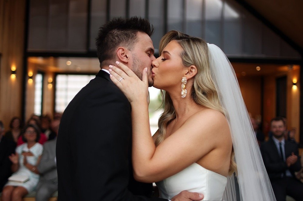 A bride and groom share a kiss at their wedding ceremony, with wedding guests clapping and smiling in the background.