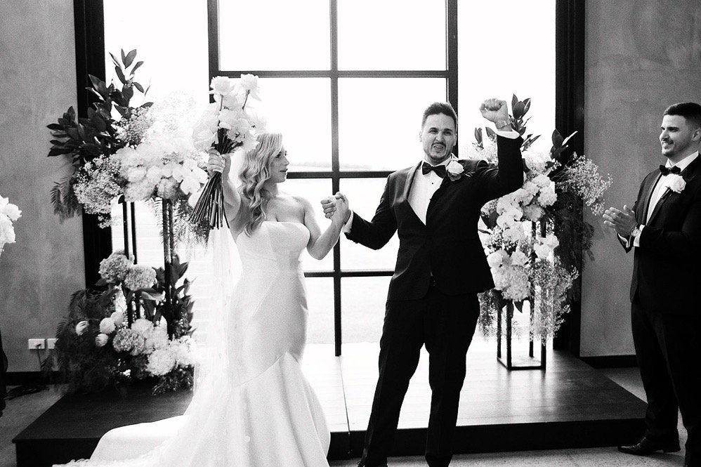 Black and white photo of a wedding ceremony showing a bride and groom, with the bride holding a bouquet, standing in front of a floral arch, with the groom celebrating, and a man clapping nearby.
