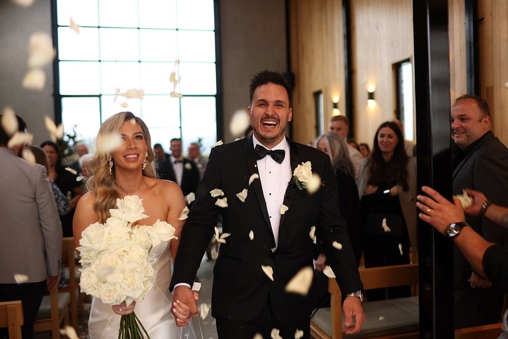 A newlywed couple holds hands and smiles as they walk through falling flower petals in a wedding ceremony, inside a decorated church or wedding venue with guests in the background.