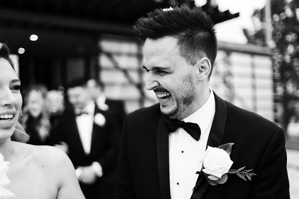 Black and white photo of a smiling woman and a man in a tuxedo with a boutonniere, at a formal outdoor event.