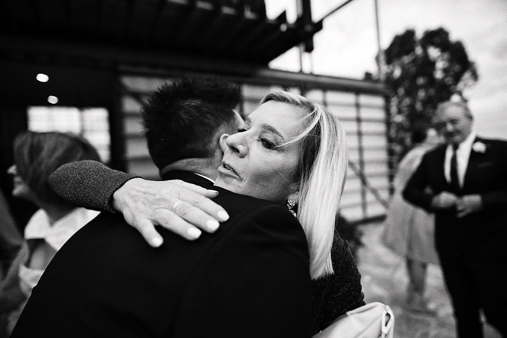 A woman embracing a man in a hug at an outdoor event, with people in formal attire in the background.