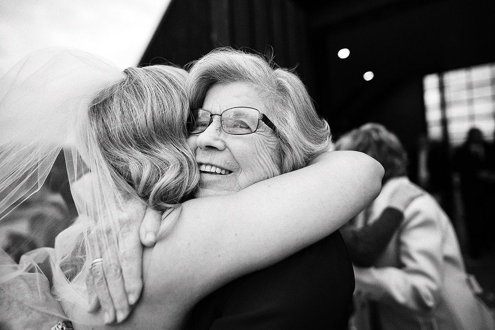 Elderly woman in glasses hugging a woman in a wedding veil, both smiling, in a joyful embrace at a celebration.