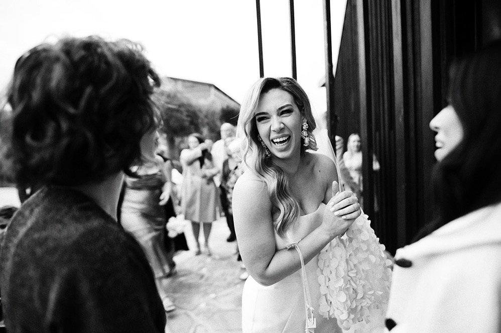 A woman in a wedding dress laughing and holding her hands together, talking to two women in a group outdoor setting.