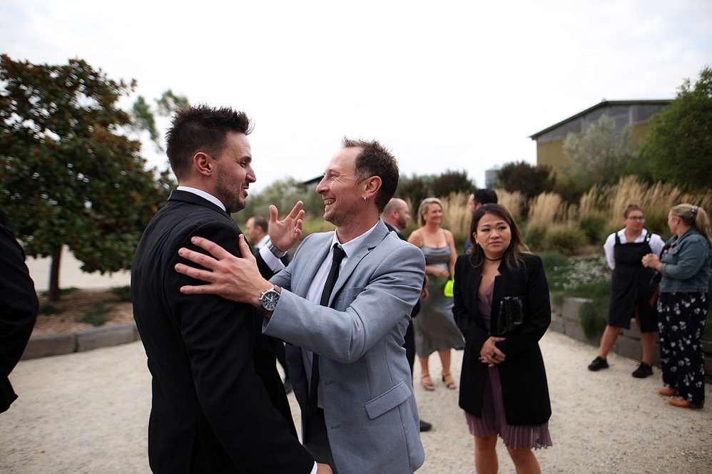 Two men dressed in suits embracing and smiling at each other outdoors, with several women in the background nearby.