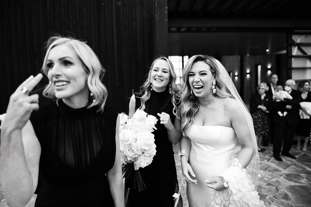 Three women at a wedding celebration, with the bride wearing a wedding dress and veil, and the two women in black dresses smiling and taking a selfie.