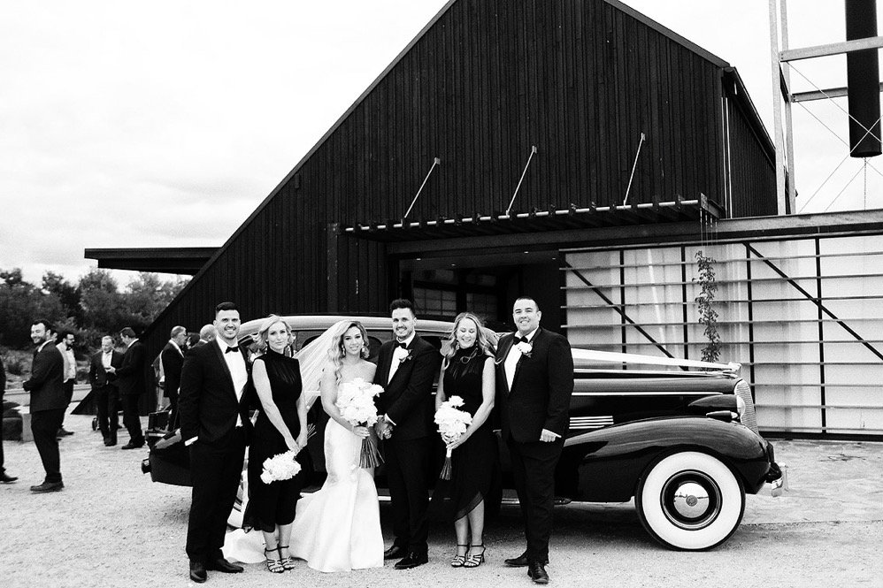 People dressed in formal attire at a wedding, standing in front of a vintage car in front of a barn-like building, with some guests in the background.