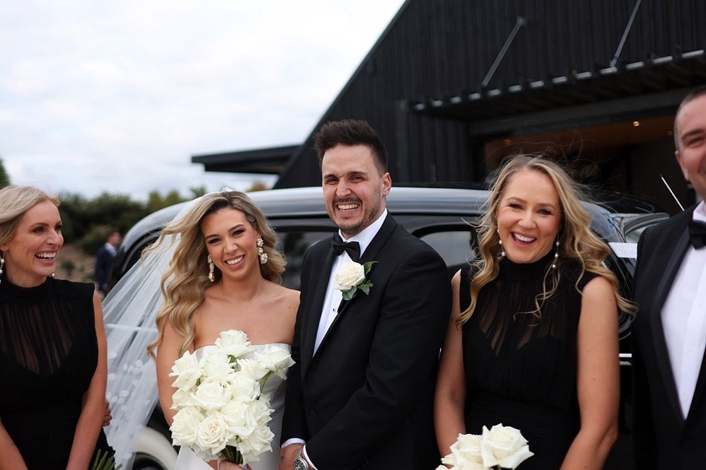 Group of people dressed in formal wedding attire, standing in front of a black vehicle near a modern building, smiling and celebrating outdoors.