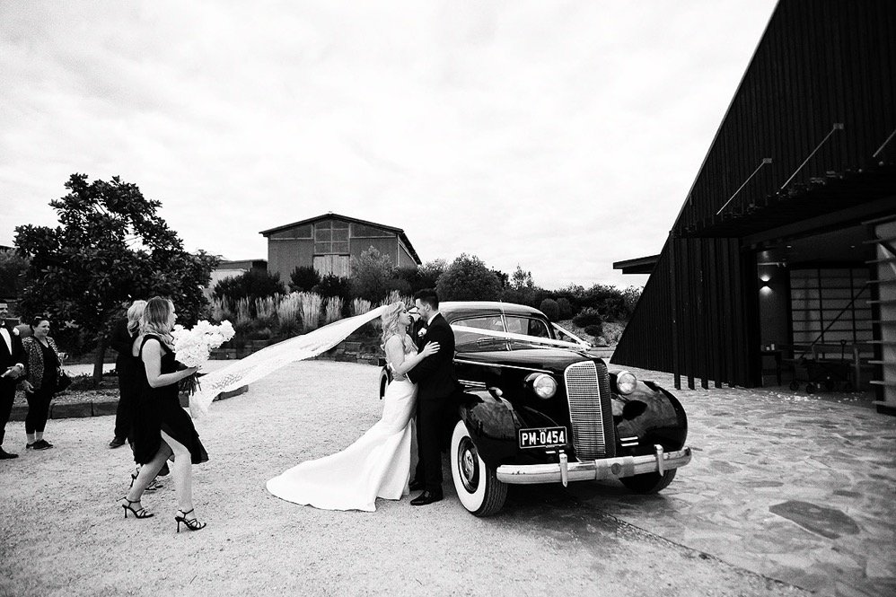 Bride and groom sharing a kiss next to a vintage black car during their wedding, with a bridesmaid holding a bouquet and guests in the background.