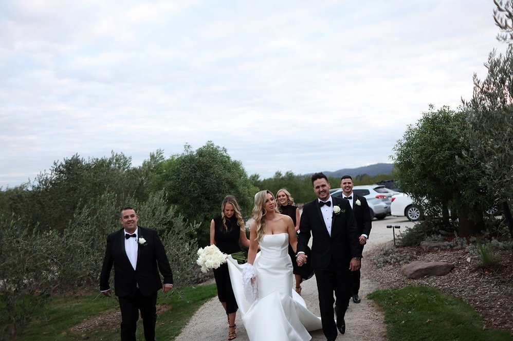 A bride and groom walking outdoors with wedding party members, surrounded by greenery and parked cars.